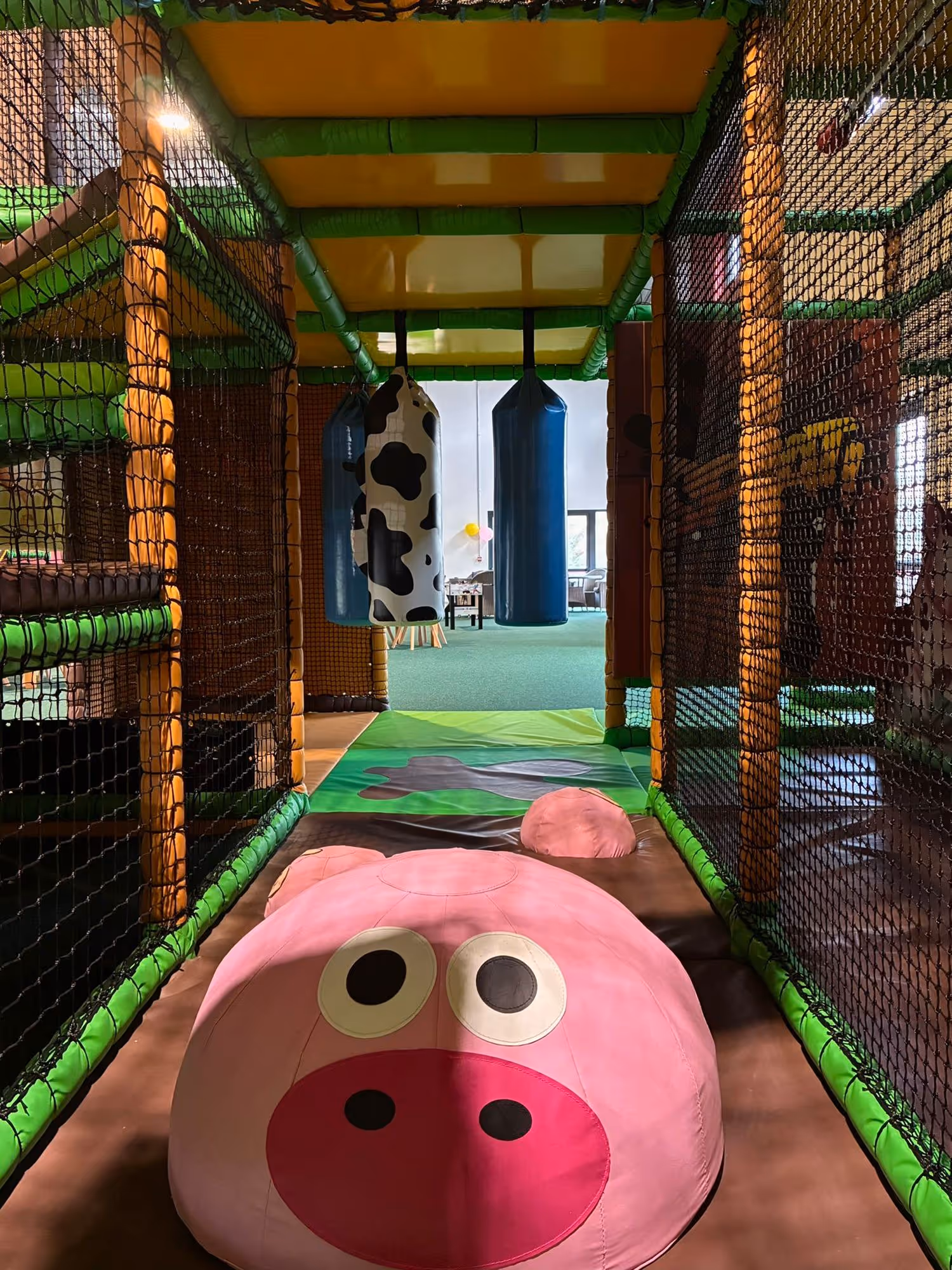 Indoor children's play area with padded floor shaped like a pink pig face, netted sides, and hanging punching bags, one with cow print.