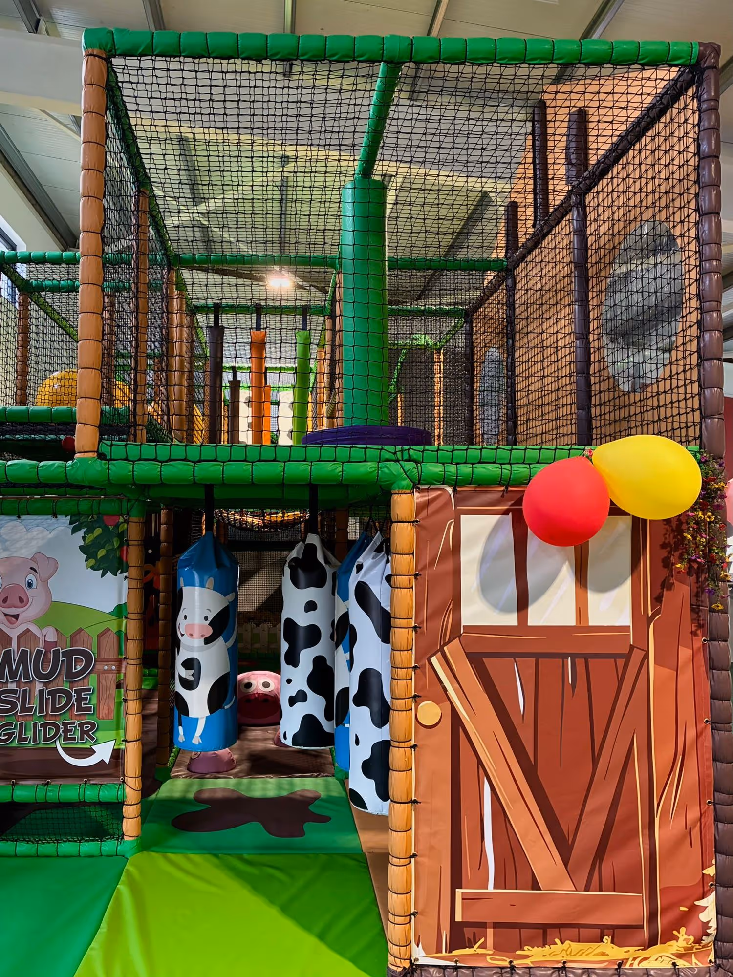Indoor children's play area with green padded structure, hanging cow-print shapes, a barn-themed entrance, and colorful balloons.