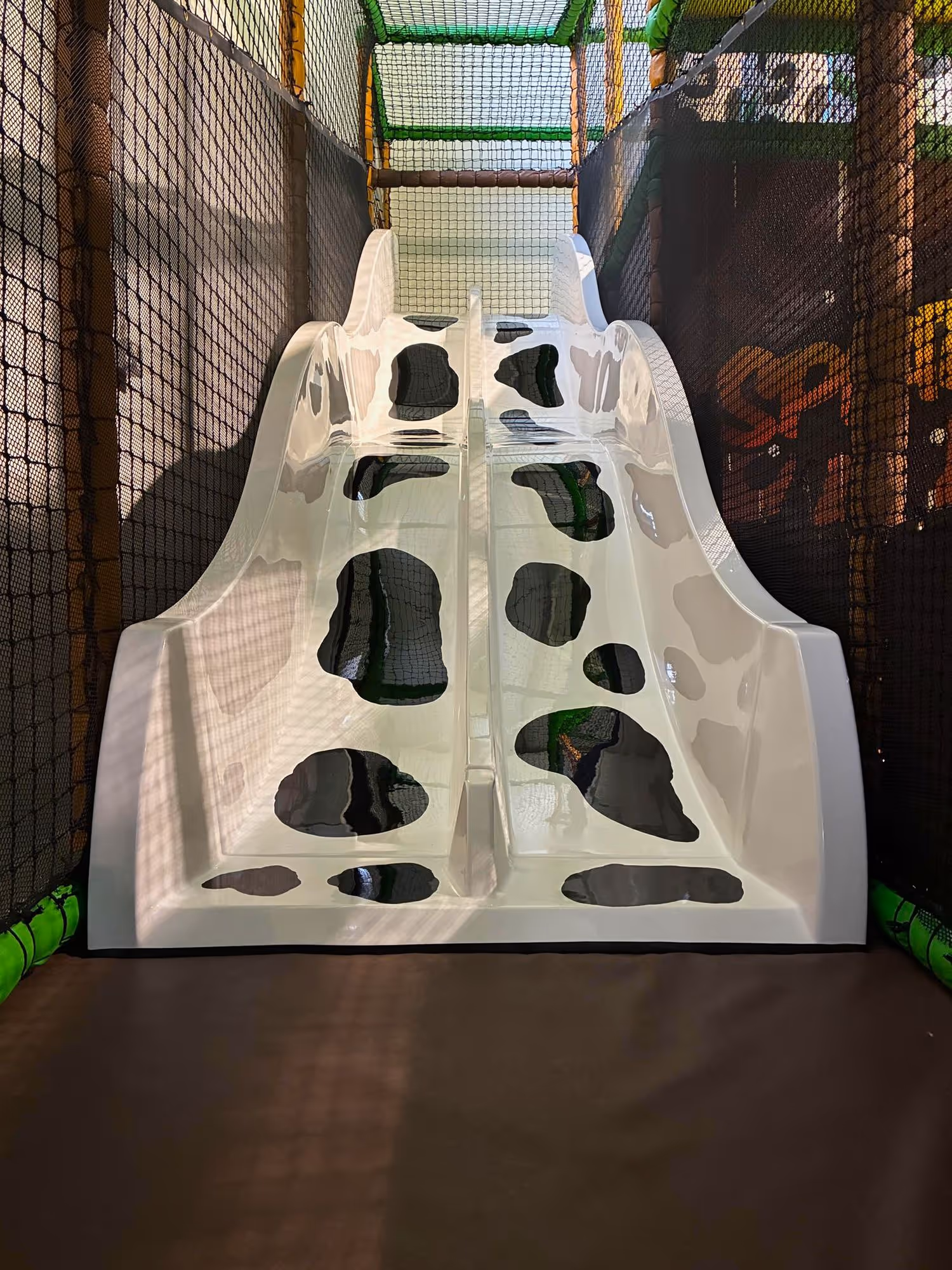 Double white children’s slide, decorated with black cow spots, in an enclosed play structure surrounded by safety netting.