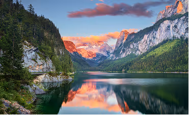 Serene lake reflecting pine trees and rocky mountains under a colorful sunset sky.