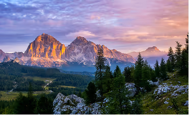 Sunset over rocky mountains with forested hills and a winding road in the valley below.