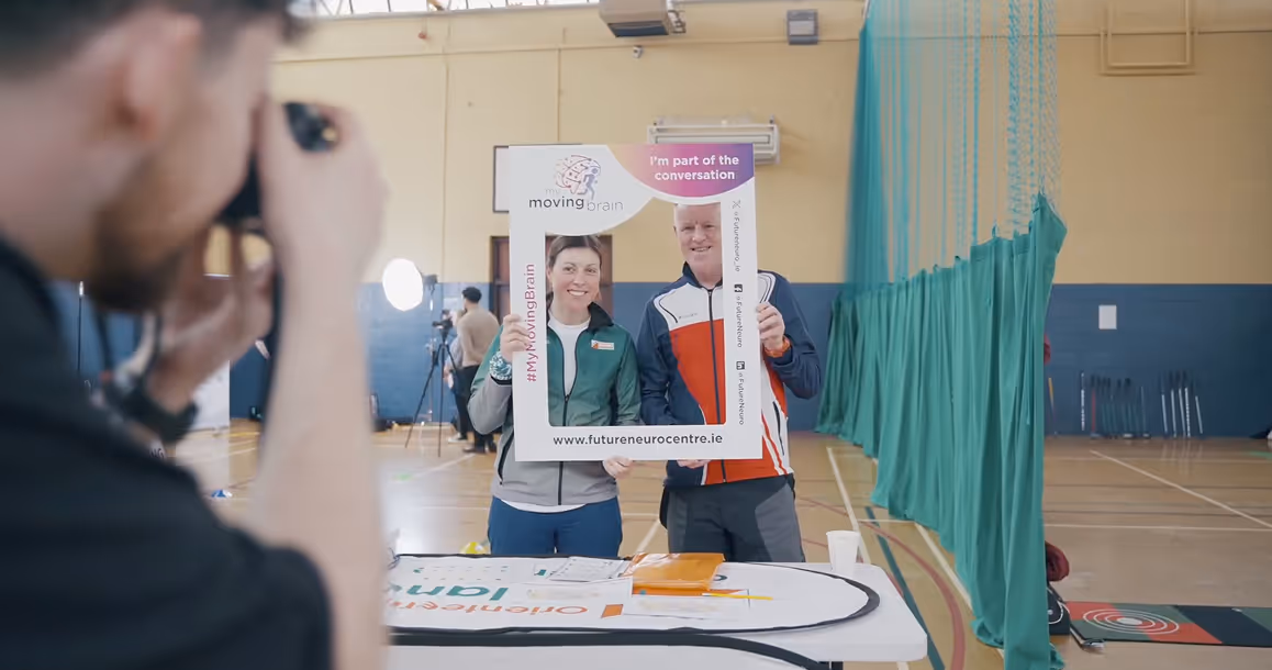 Two people smiling and holding a photo frame prop with 'moving brain' logo indoors.