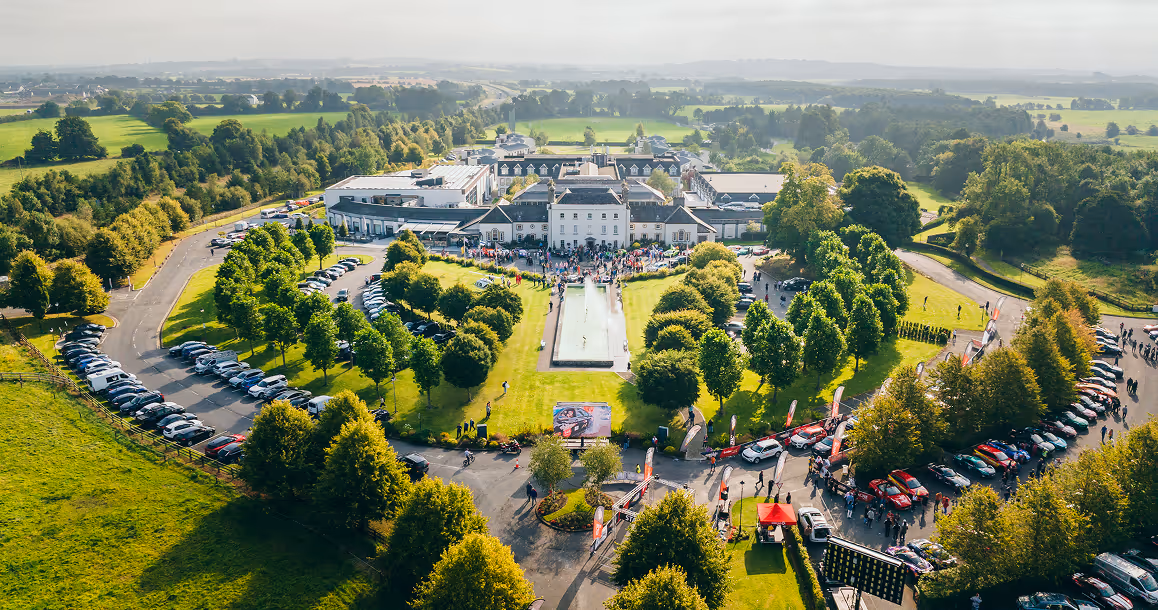 Aerial view of a large estate with a central building, fountain, trees, parked cars, and people outdoors.