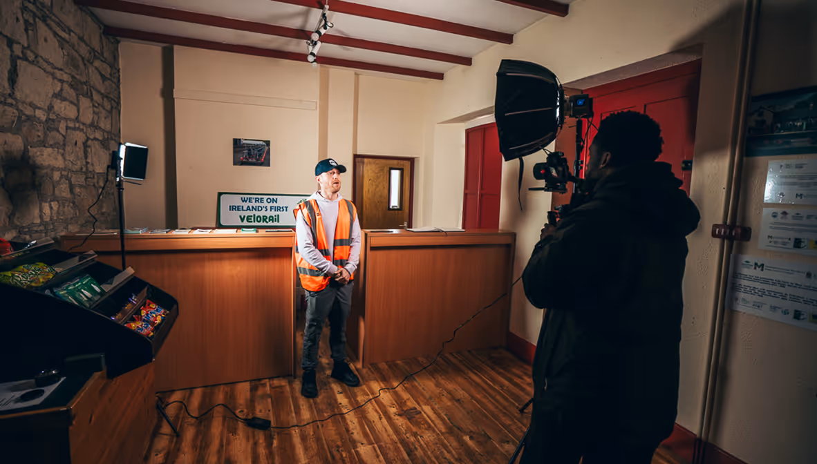 Man in orange safety vest and cap standing behind a wooden counter while being filmed indoors.