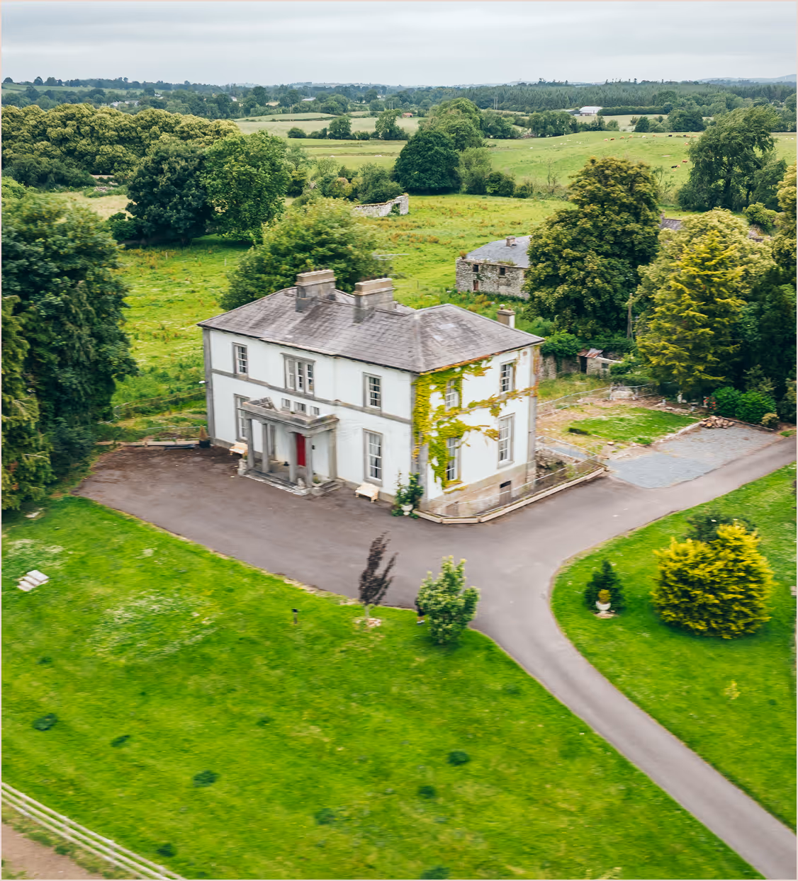 A two-story white house with greenery and trees around, surrounded by lush green fields.