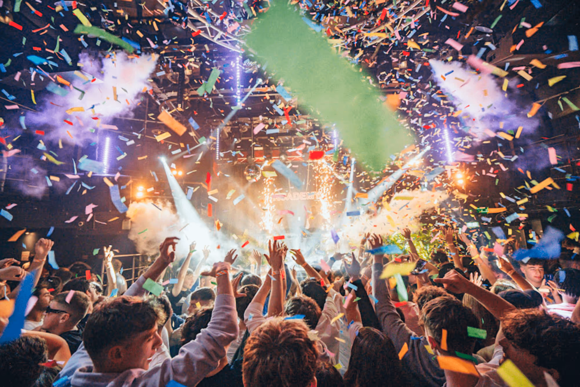 Crowd at a concert with colorful confetti and bright stage lights creating a festive atmosphere.