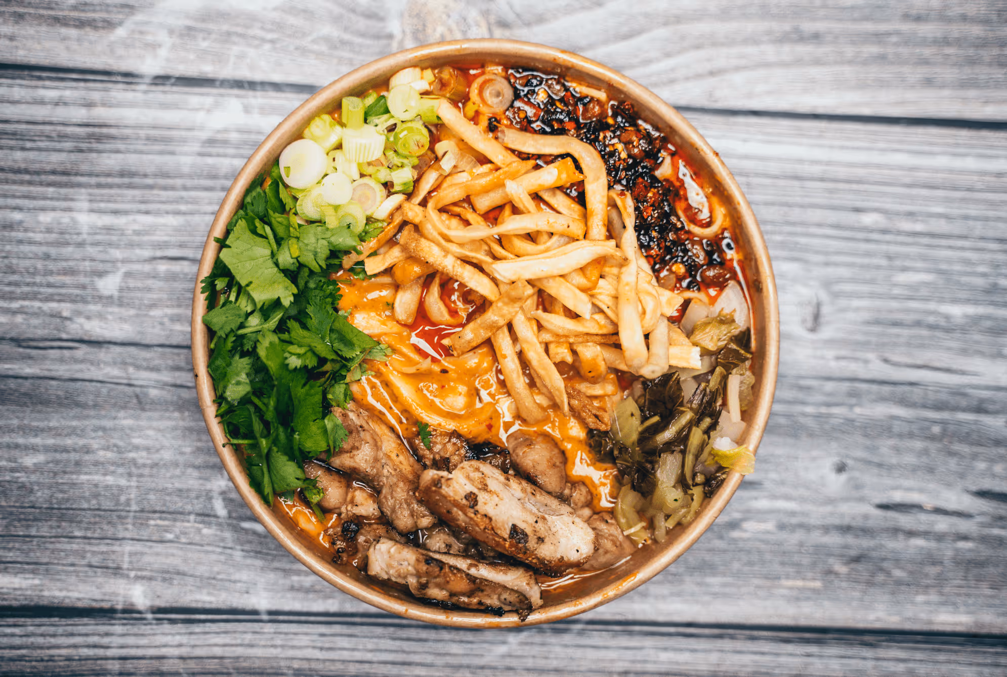 Bowl of ramen with grilled chicken, fries, chopped scallions, greens, spicy sauce, and chili oil on wood table.