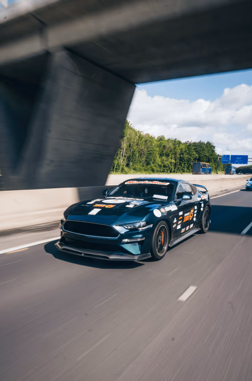 Blue racing car with sponsor decals driving on a highway under a concrete overpass.