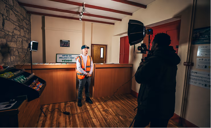 Man in orange safety vest being filmed indoors with professional camera and lighting equipment.