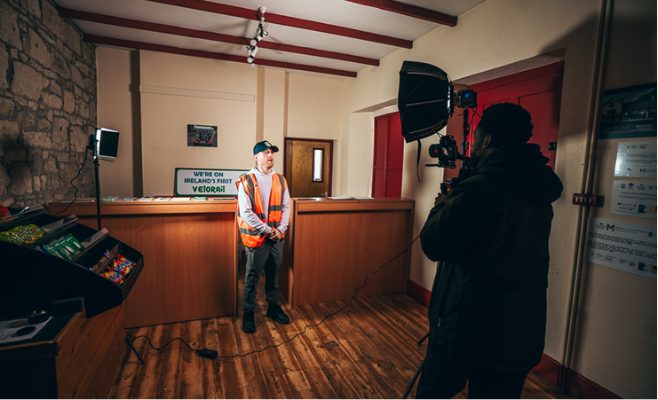 Man in orange safety vest being filmed indoors with professional camera and lighting equipment.