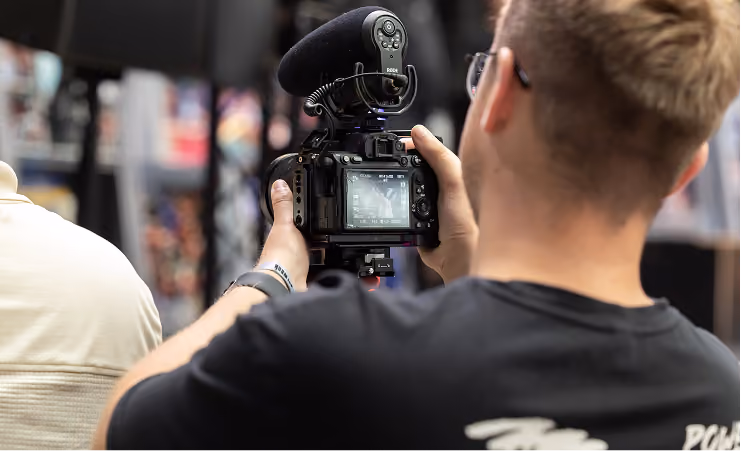 Man with glasses filming with a camera that has an attached Rode microphone indoors.