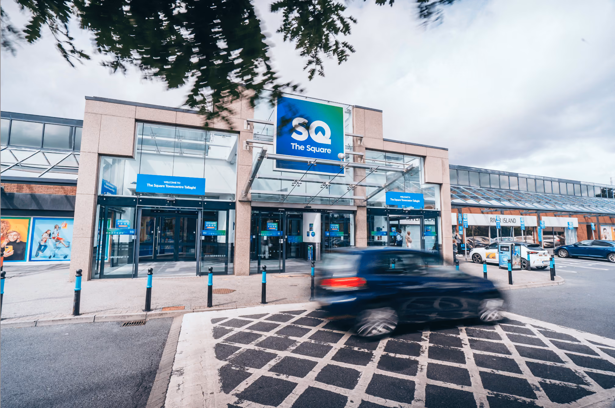Entrance of The Square shopping center with a blurred black car driving past the front.