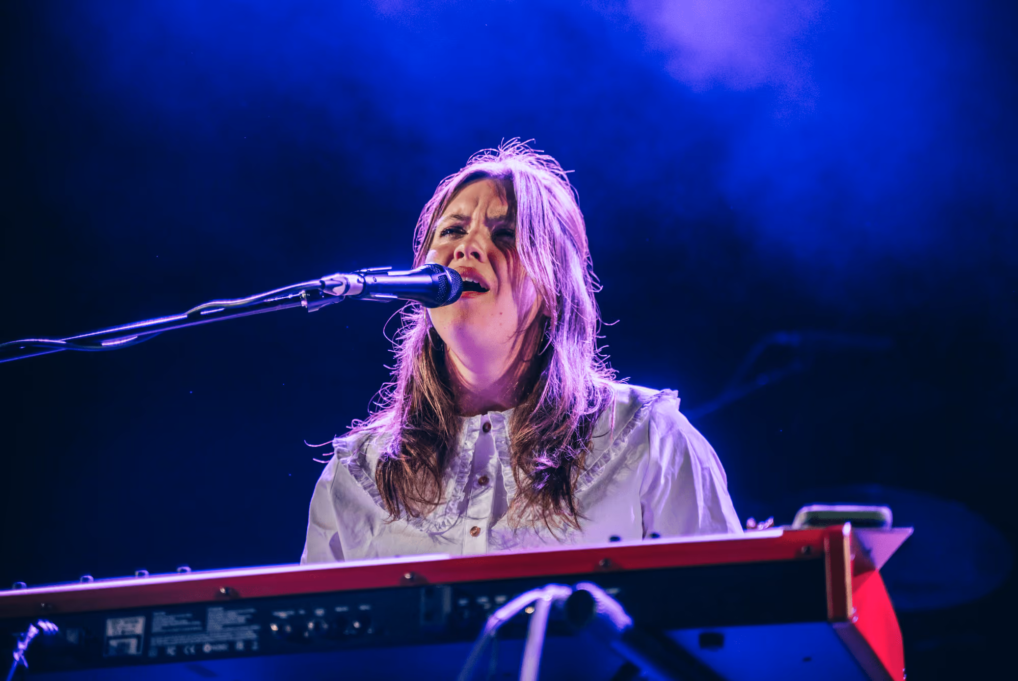Female musician singing into a microphone while playing a keyboard on stage with blue lighting.