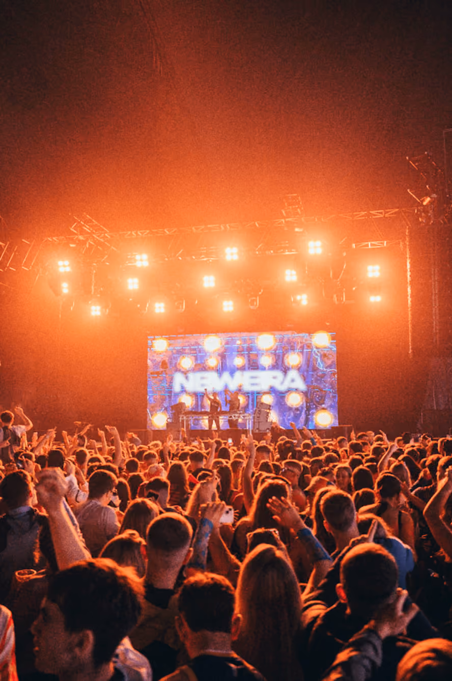 Large crowd at a concert with raised hands facing a brightly lit stage with NewEra text on screen.