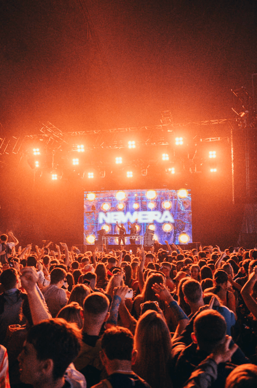 Large crowd at a concert with raised hands facing a brightly lit stage with NewEra text on screen.