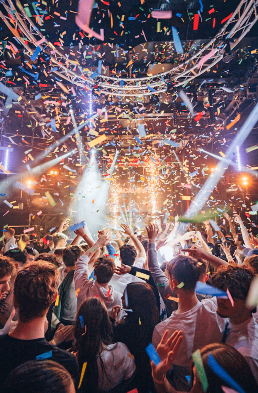 Crowd at a lively indoor party with confetti falling and bright lights on stage.