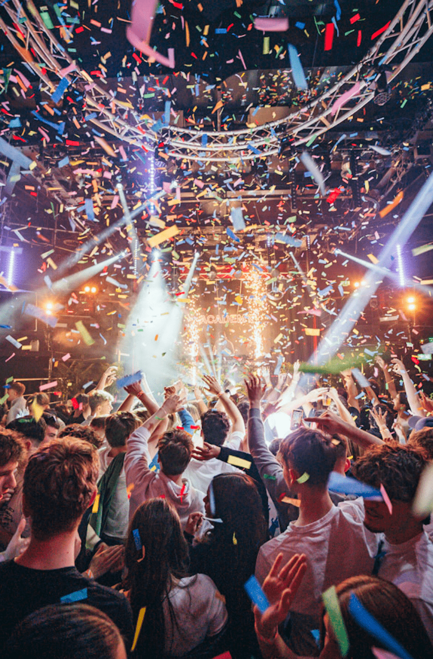 Crowd at a lively indoor party with confetti falling and bright lights on stage.