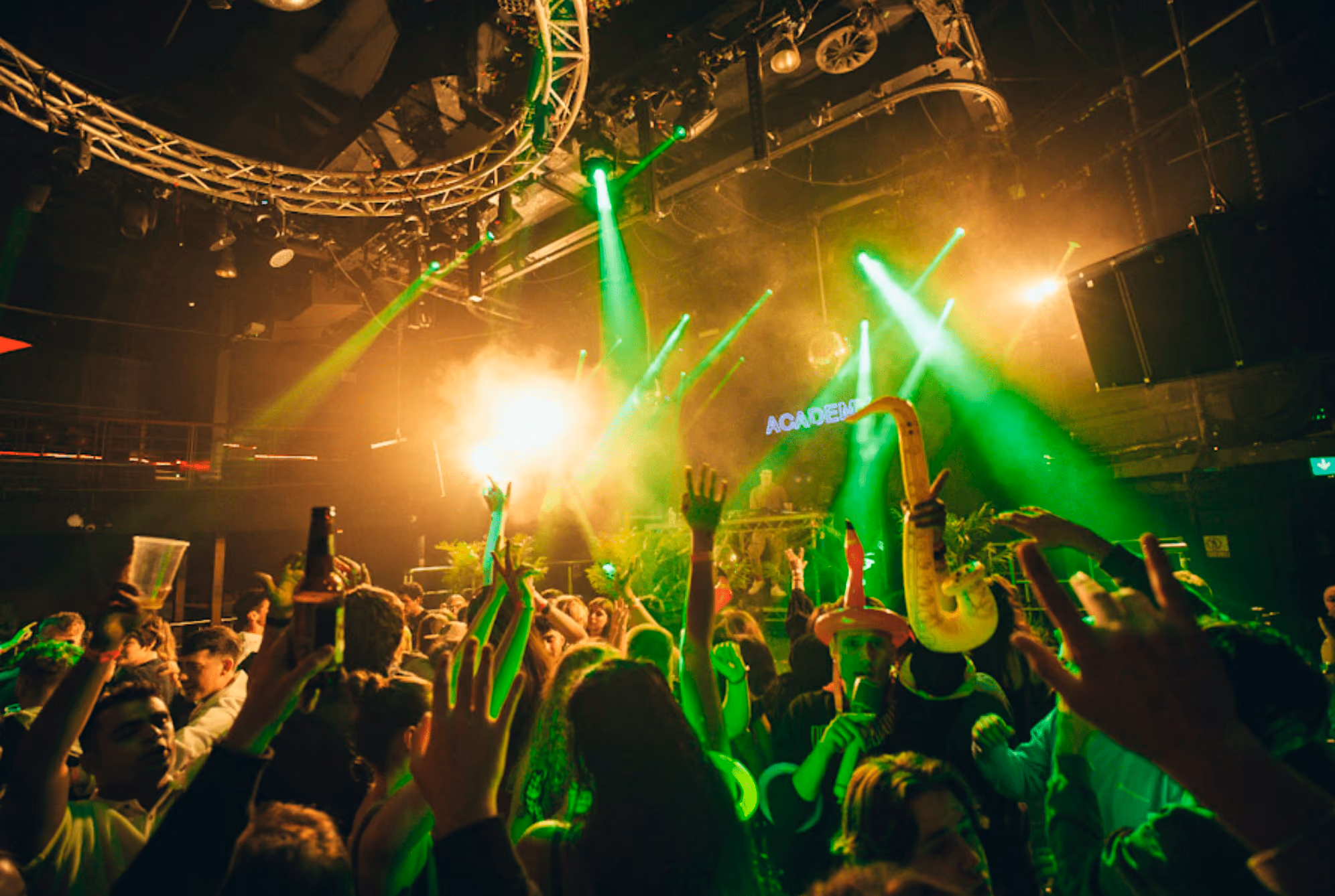 Crowd dancing and raising hands under green stage lights at a lively indoor concert or club event.