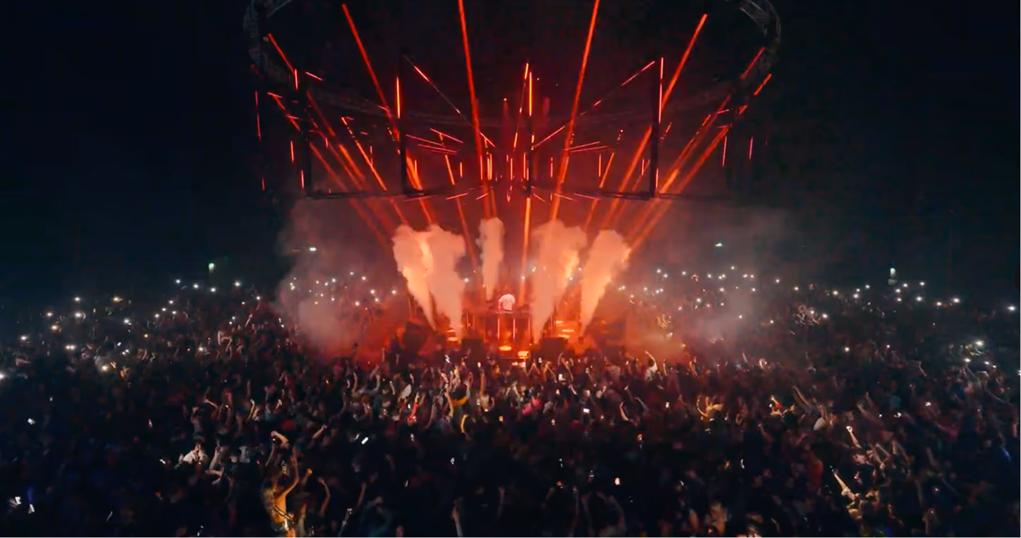 Crowd at a night concert with a DJ, red lights, smoke effects, and many phone flashlights visible.