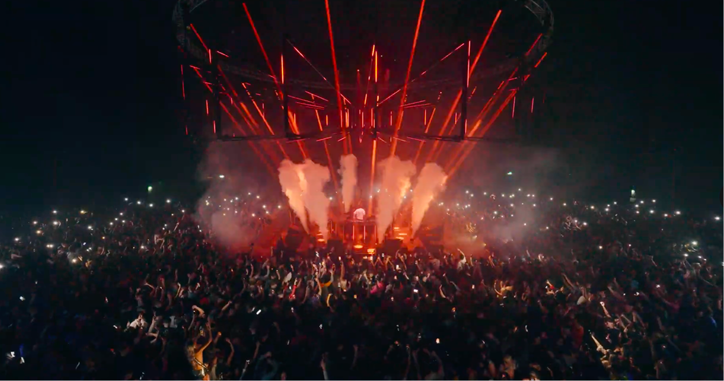 Crowd at a night concert with a DJ, red lights, smoke effects, and many phone flashlights visible.