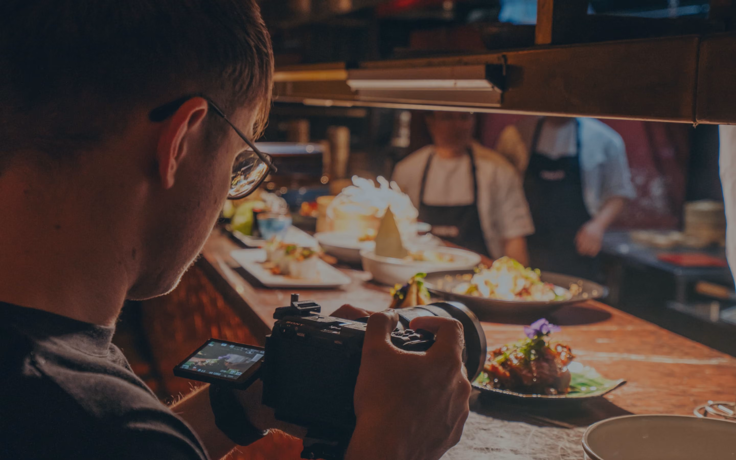Photographer in glasses capturing close-up of plated food at restaurant kitchen pass.