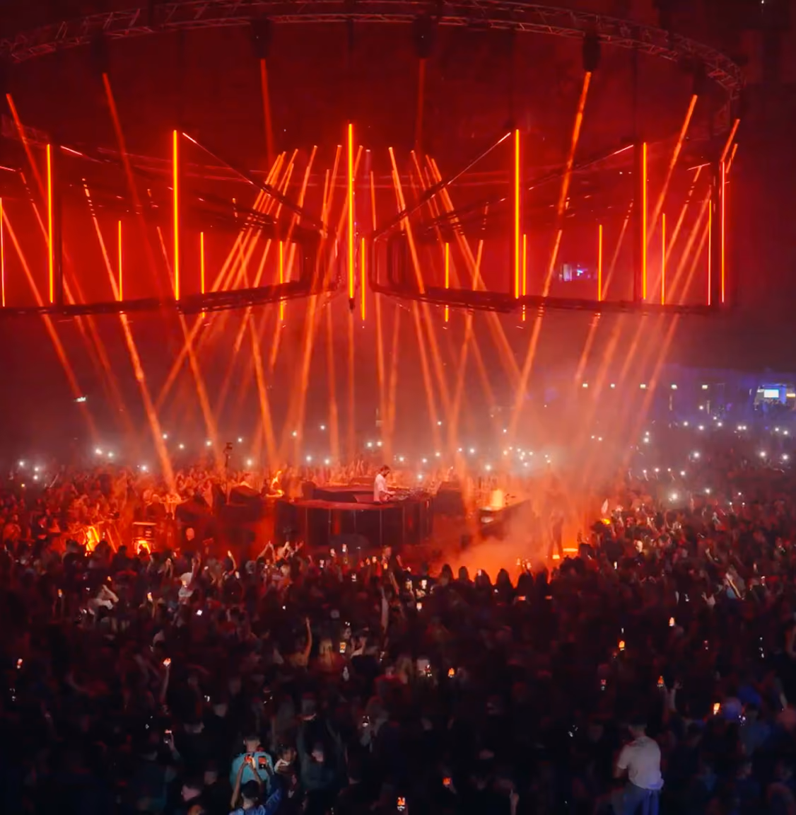 DJ performing on stage with red lights and a large crowd holding up phones at a concert.