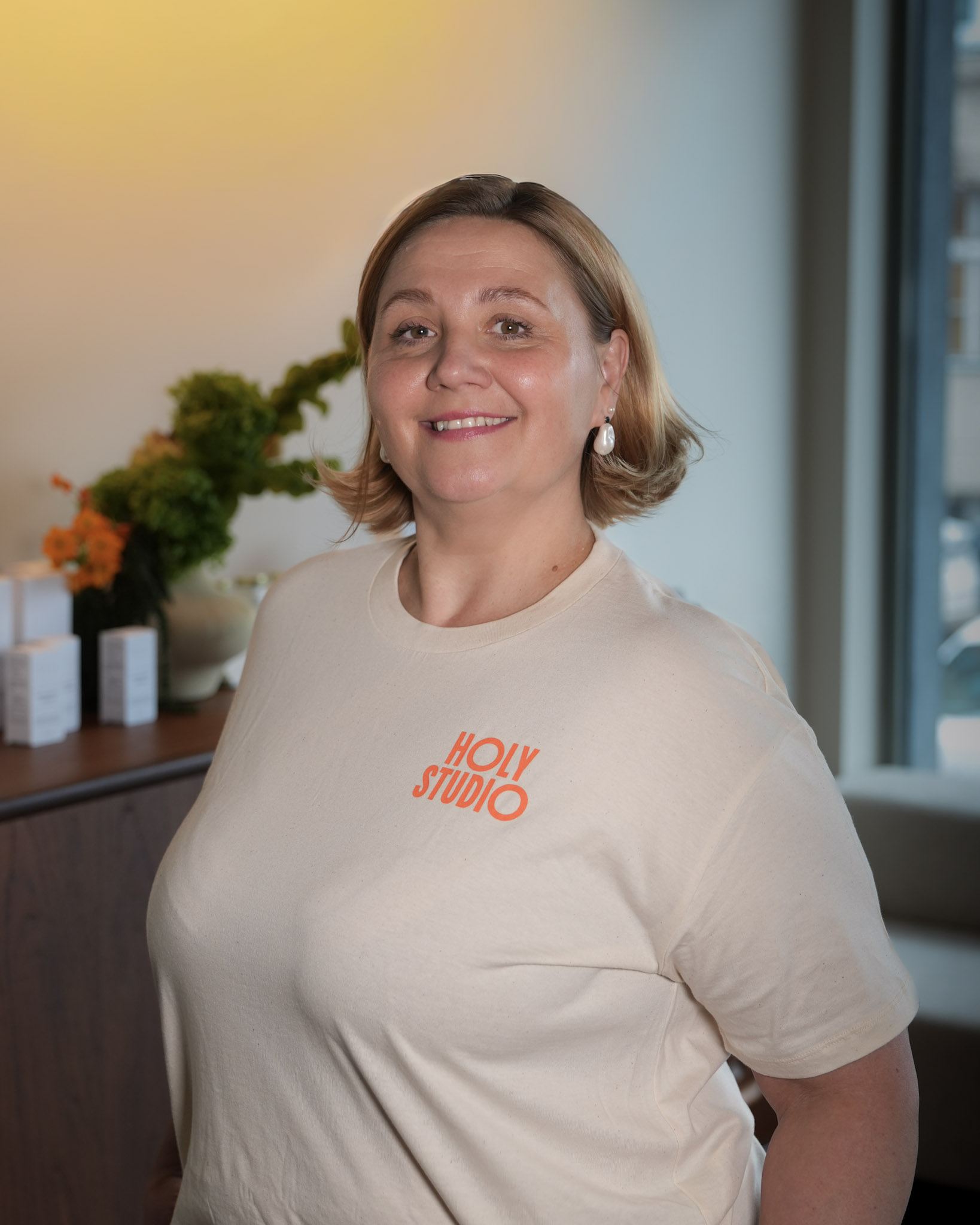 Uśmiechnięta kobieta w białej koszulce z napisem HOLY na jasnym tle.