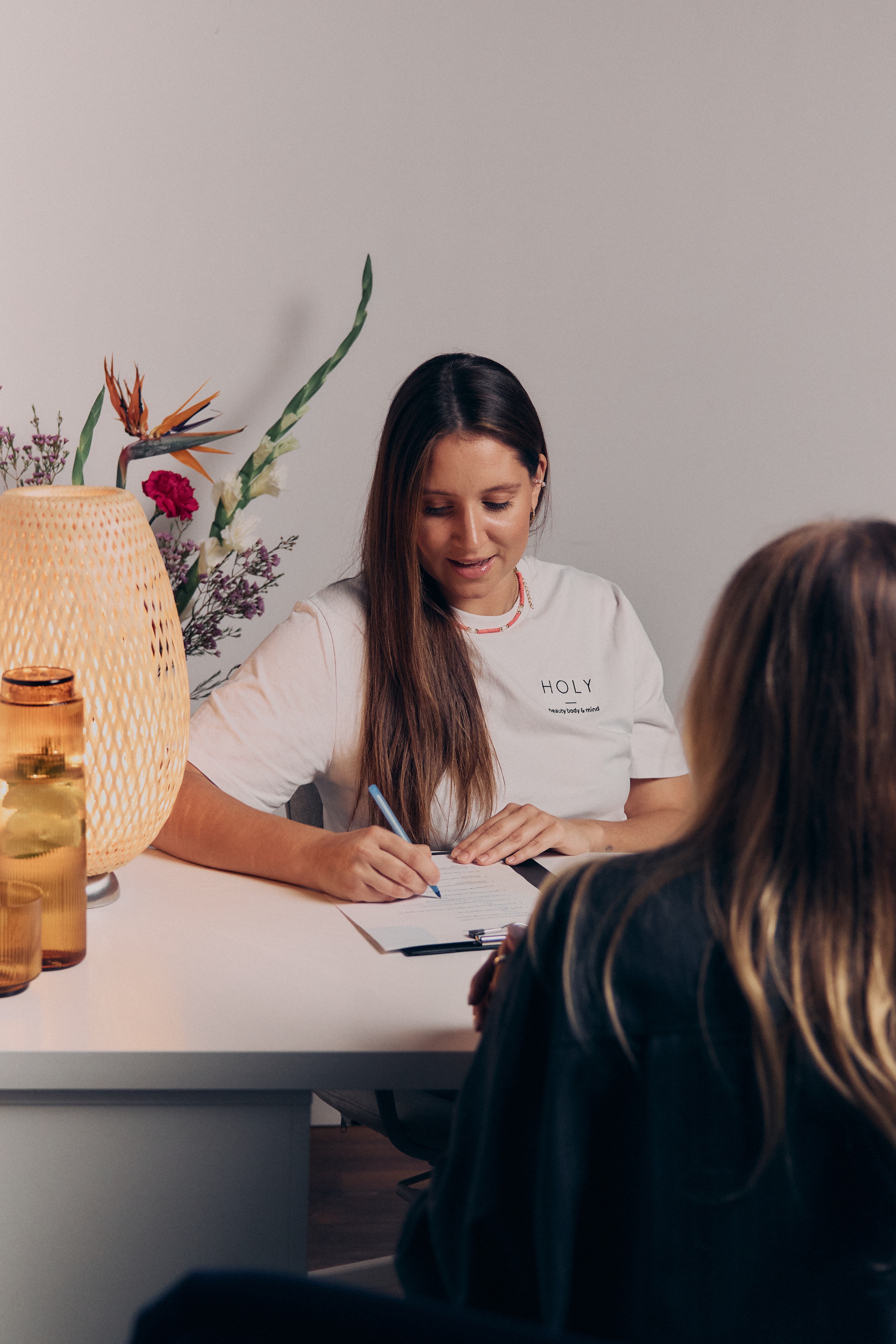 Kobieta ćwicząca rozciąganie w pozycji psa z głową w dół na macie, asystowana przez trenerkę w białej koszulce.