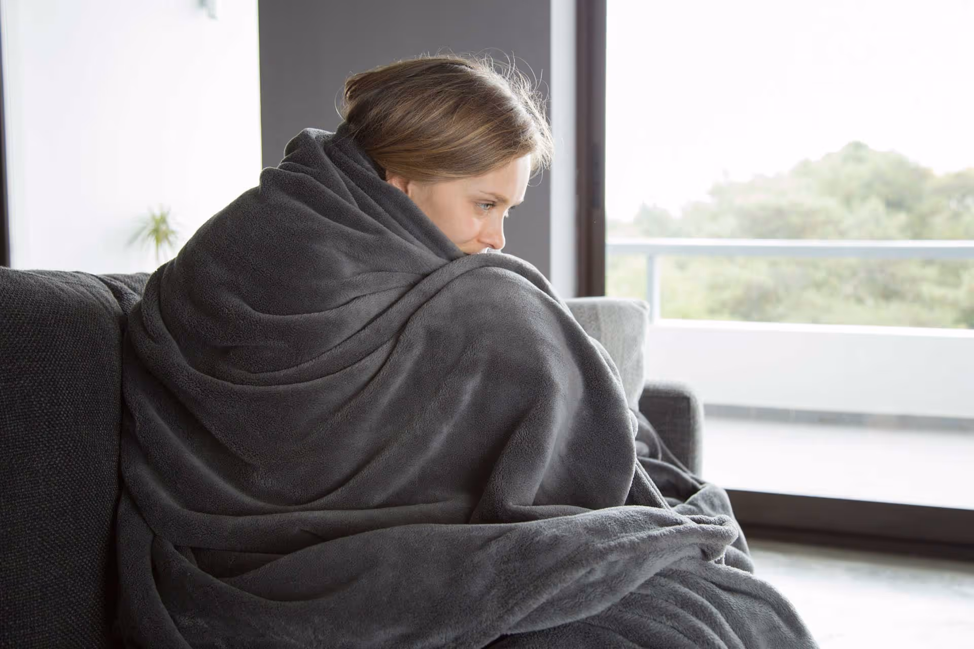 A person is wrapped in a thick grey blanket, sitting on a couch, looking thoughtful.