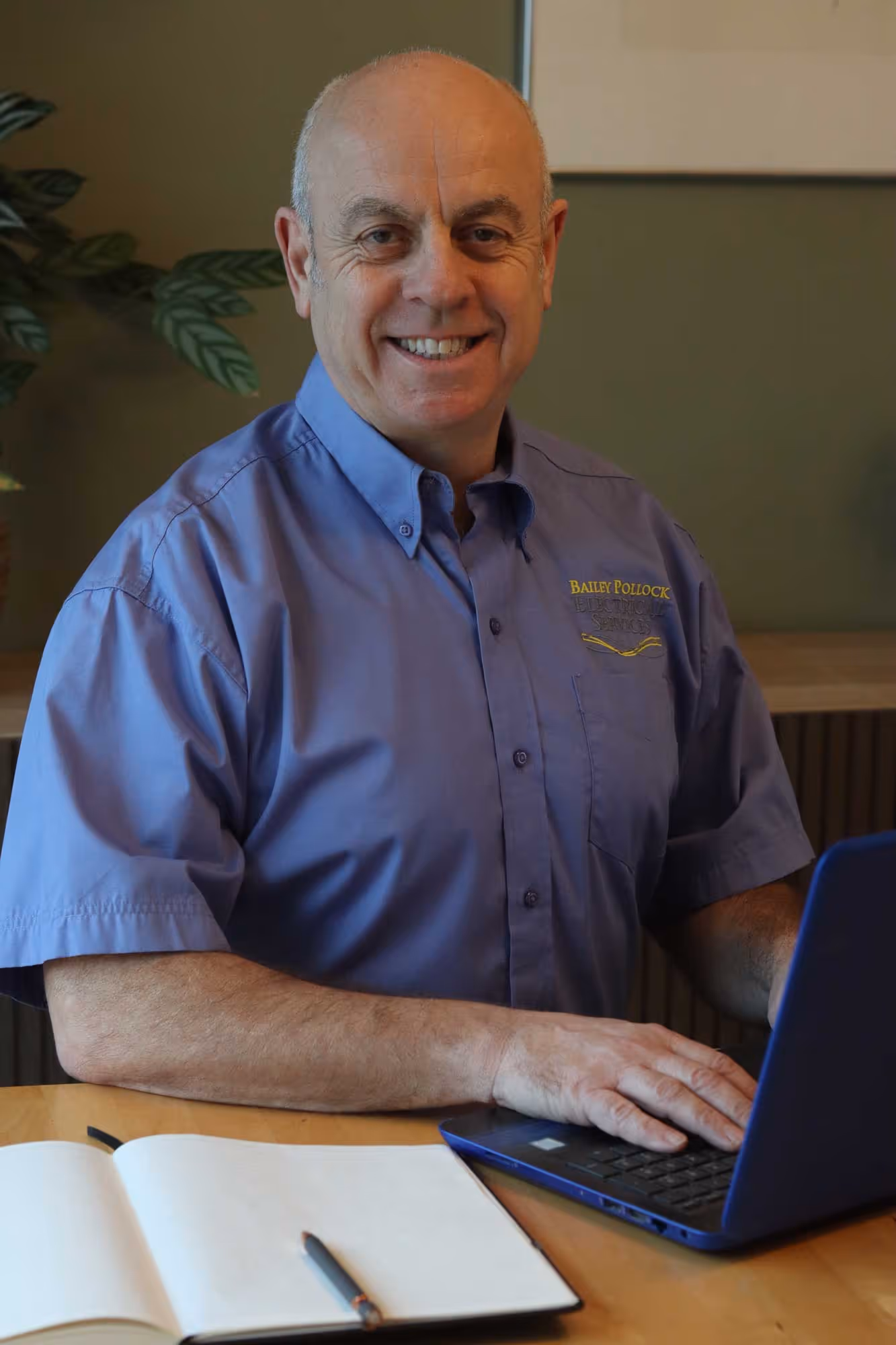 A man wearing a blue shirt is smiling and working on a laptop at a table.