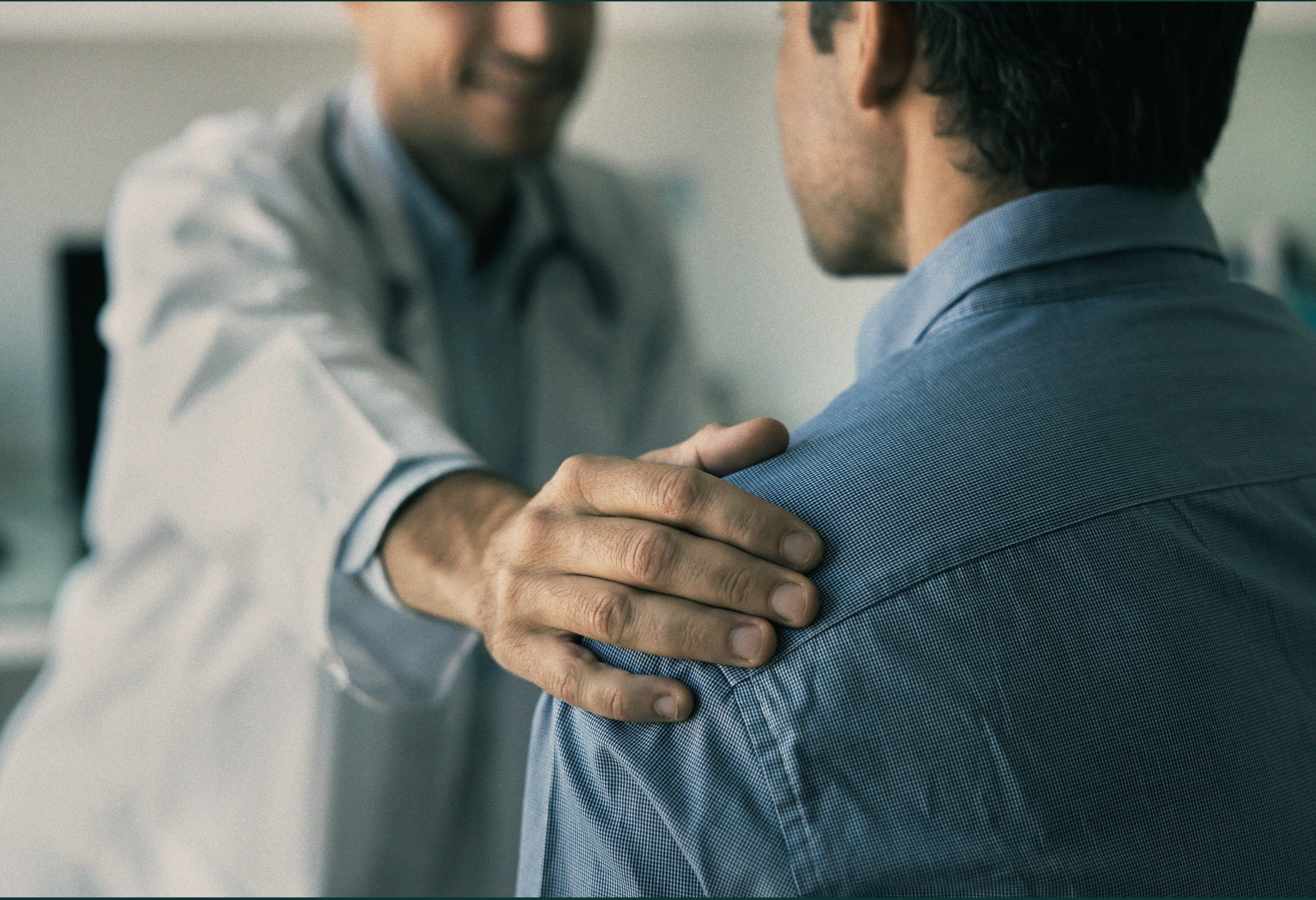 Doctor with stethoscope gently placing hand on shoulder of a man in a blue shirt.