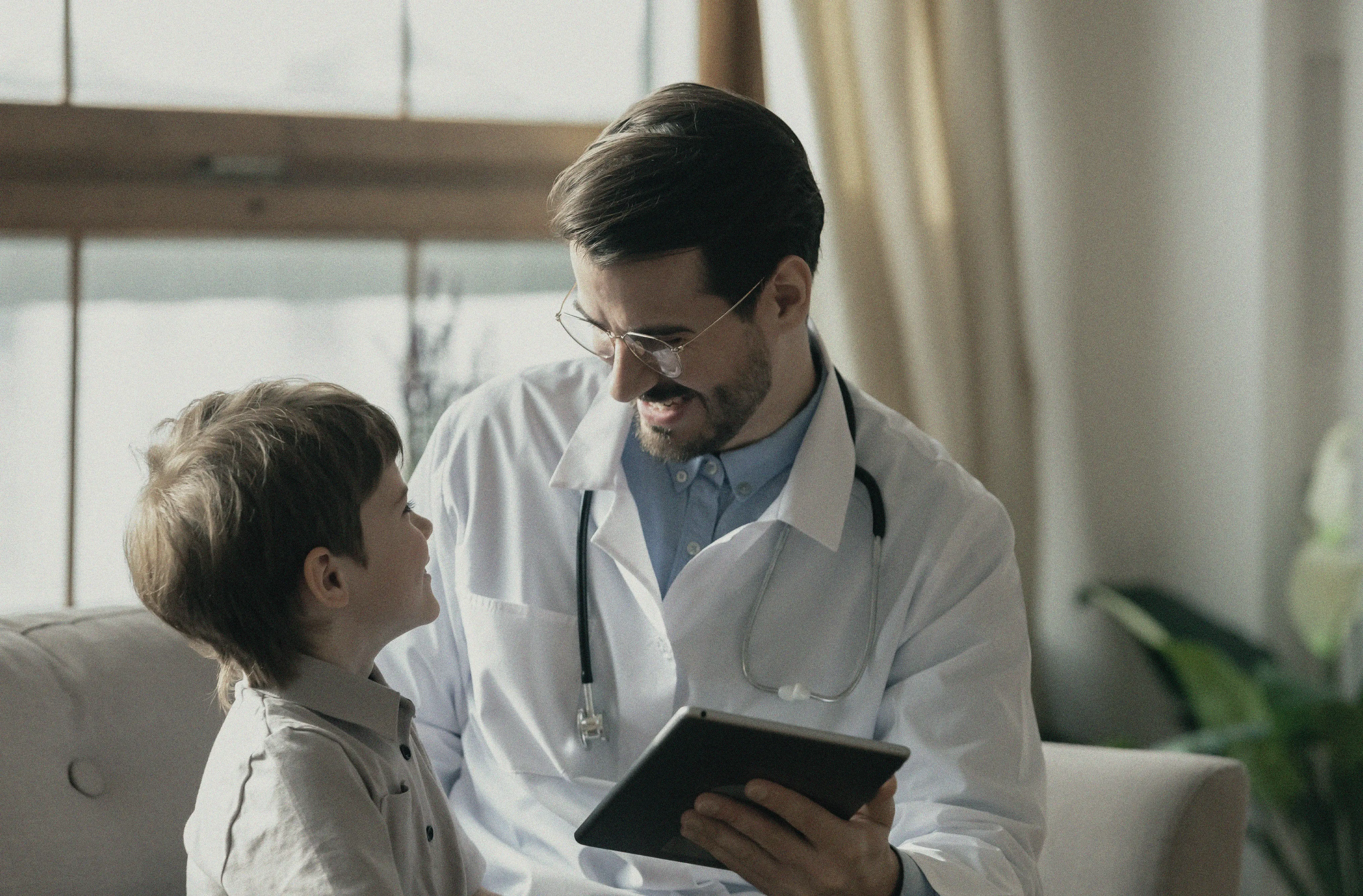 Male doctor with glasses and stethoscope smiling and showing a tablet to a young boy sitting on a couch.