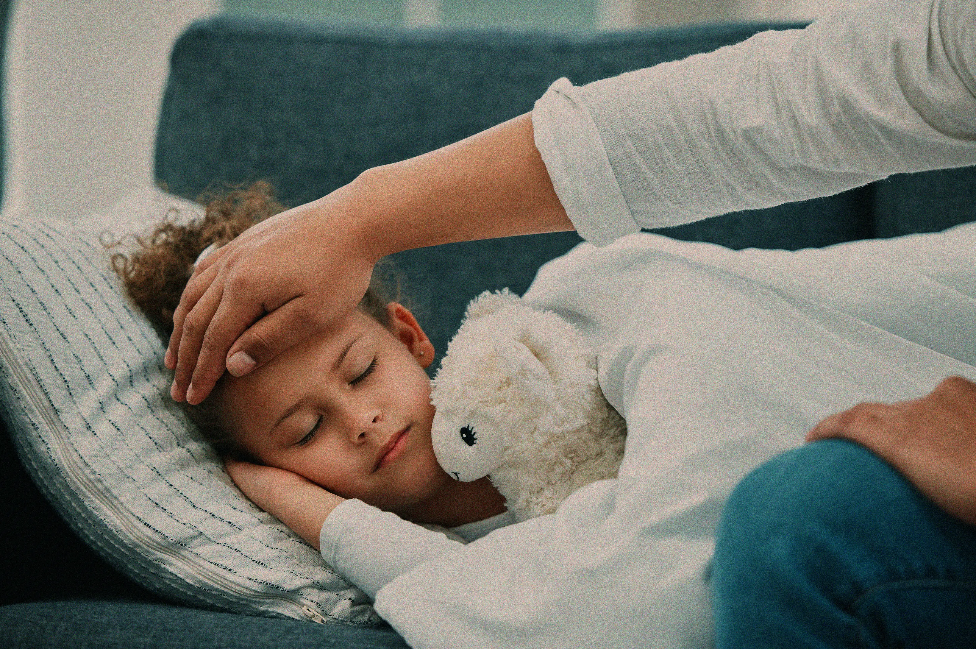 Child lying in bed with eyes closed, covered with a white blanket and holding a stuffed animal, while an adult hand touches the child's forehead.