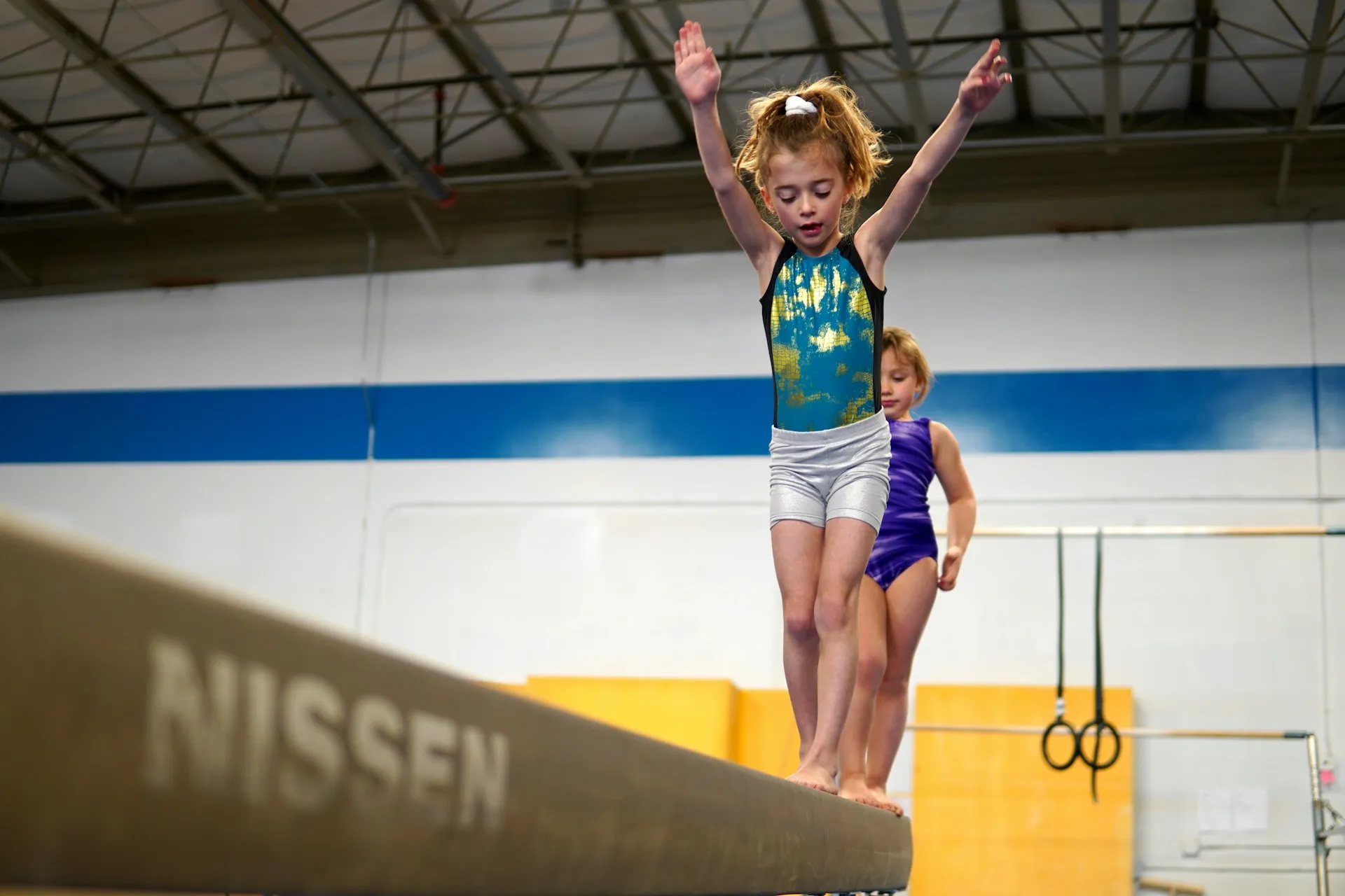 Two young girls in leotards balancing on a gymnastics beam in a gym.