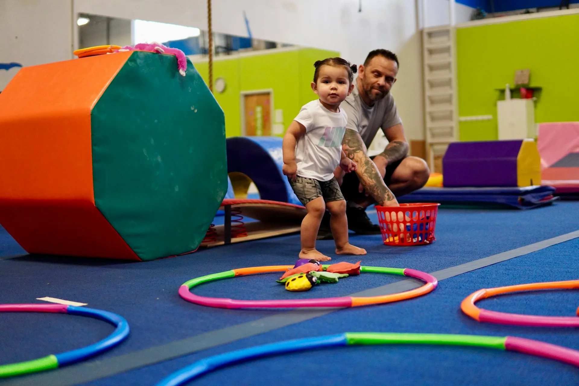 Toddler standing inside a colorful hoop on a blue gym floor, with an adult crouching nearby holding a red basket.