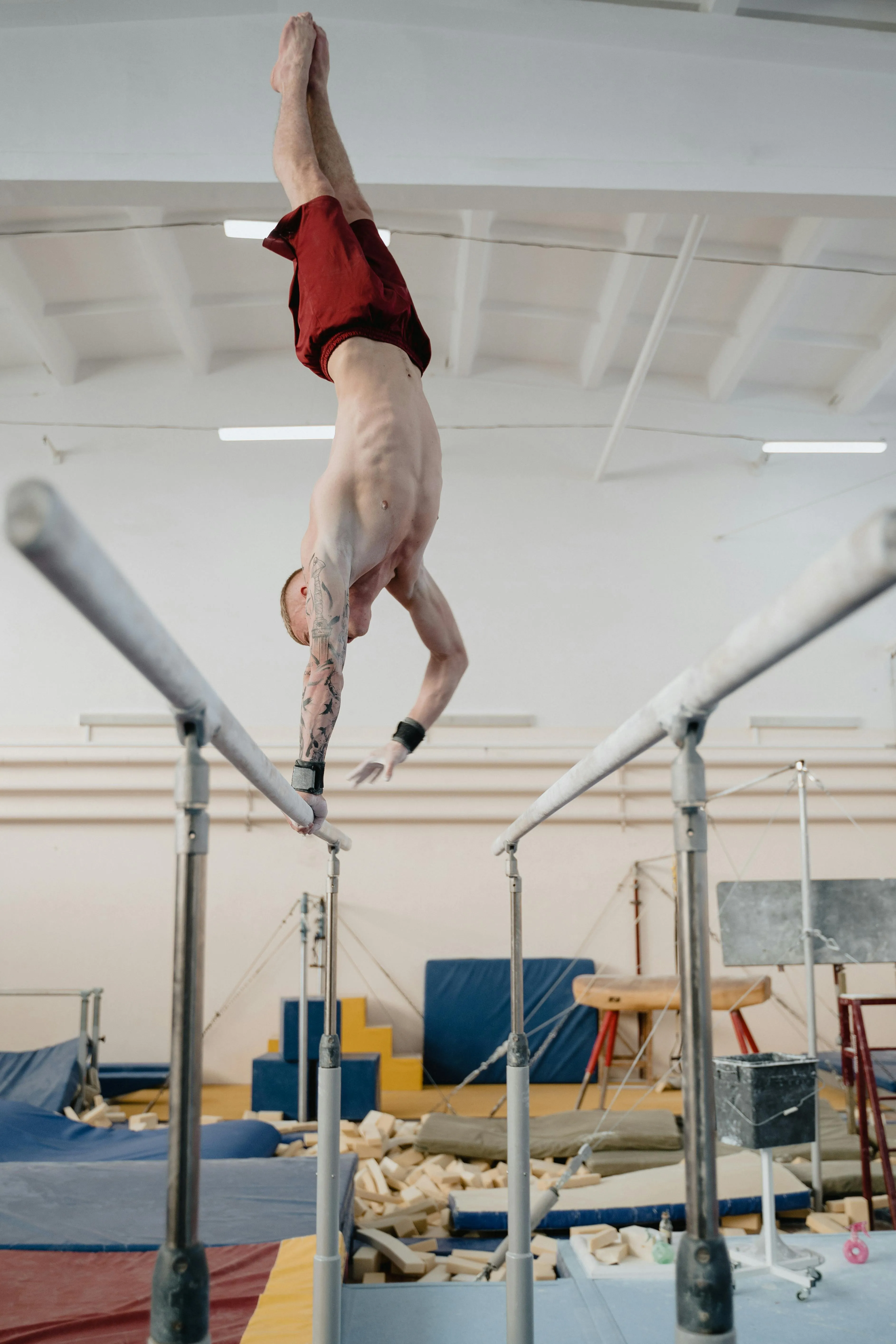 Shirtless man with tattoos performing a handstand on parallel bars in a gymnastics gym.