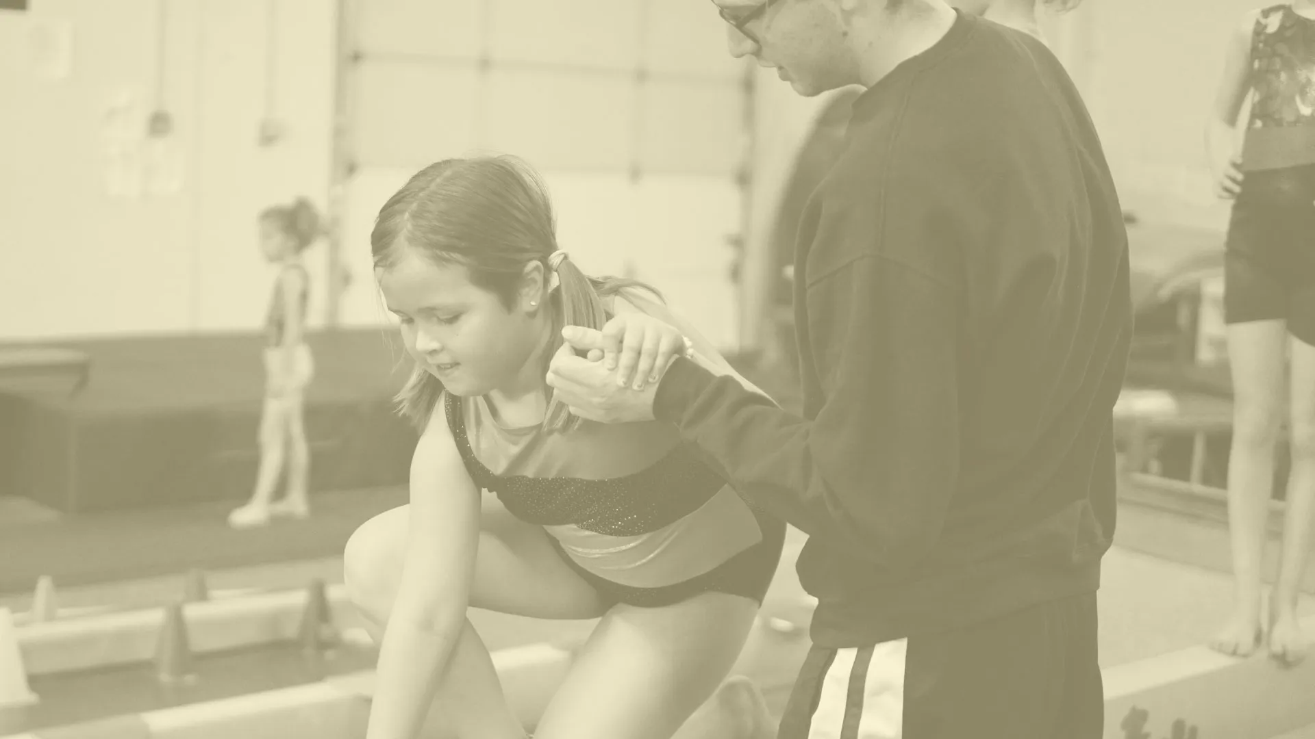 A coach assisting a young girl in gymnastics, holding her hand as she prepares to perform near a balance beam.