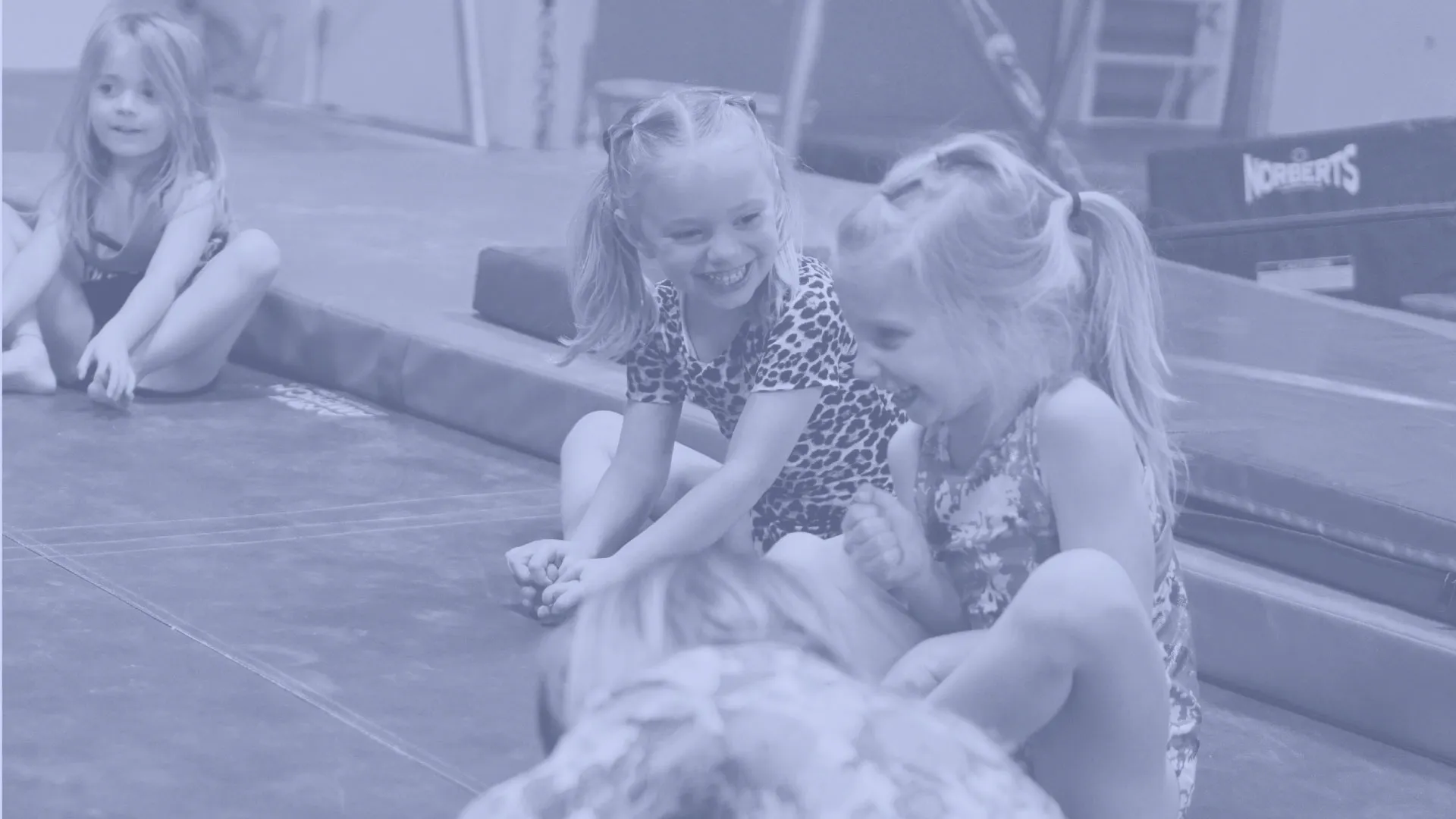 Three young girls sitting on gym mats, smiling and interacting with each other indoors.
