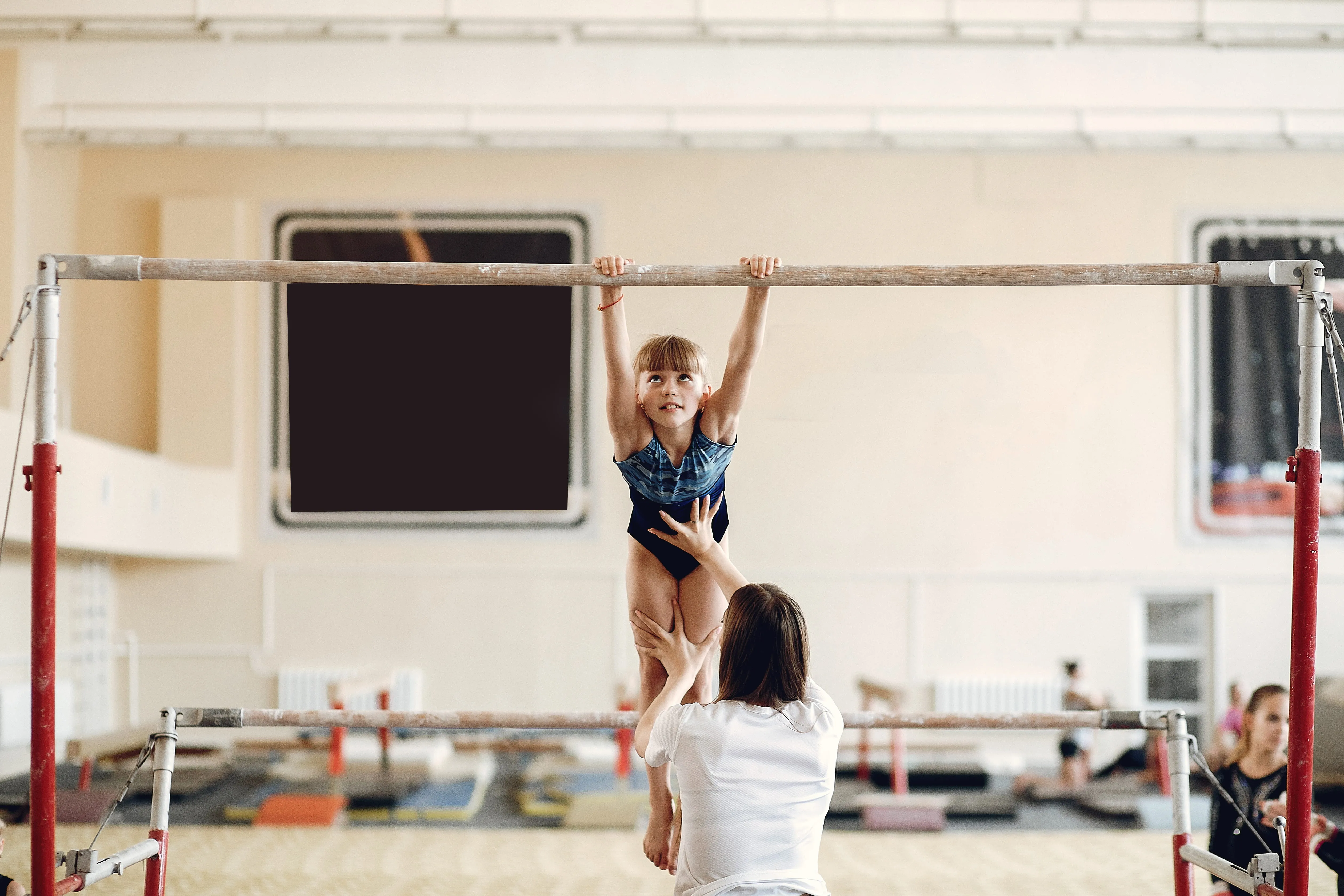 Young girl in gymnastics leotard practicing on uneven bars with coach spotting her from below in a gym.