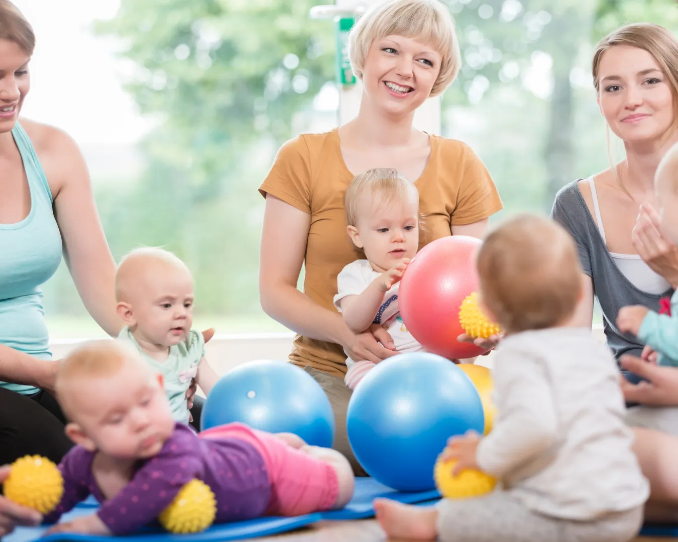 Three women sit with babies playing with colorful balls and textured toys on a blue mat in a bright room.