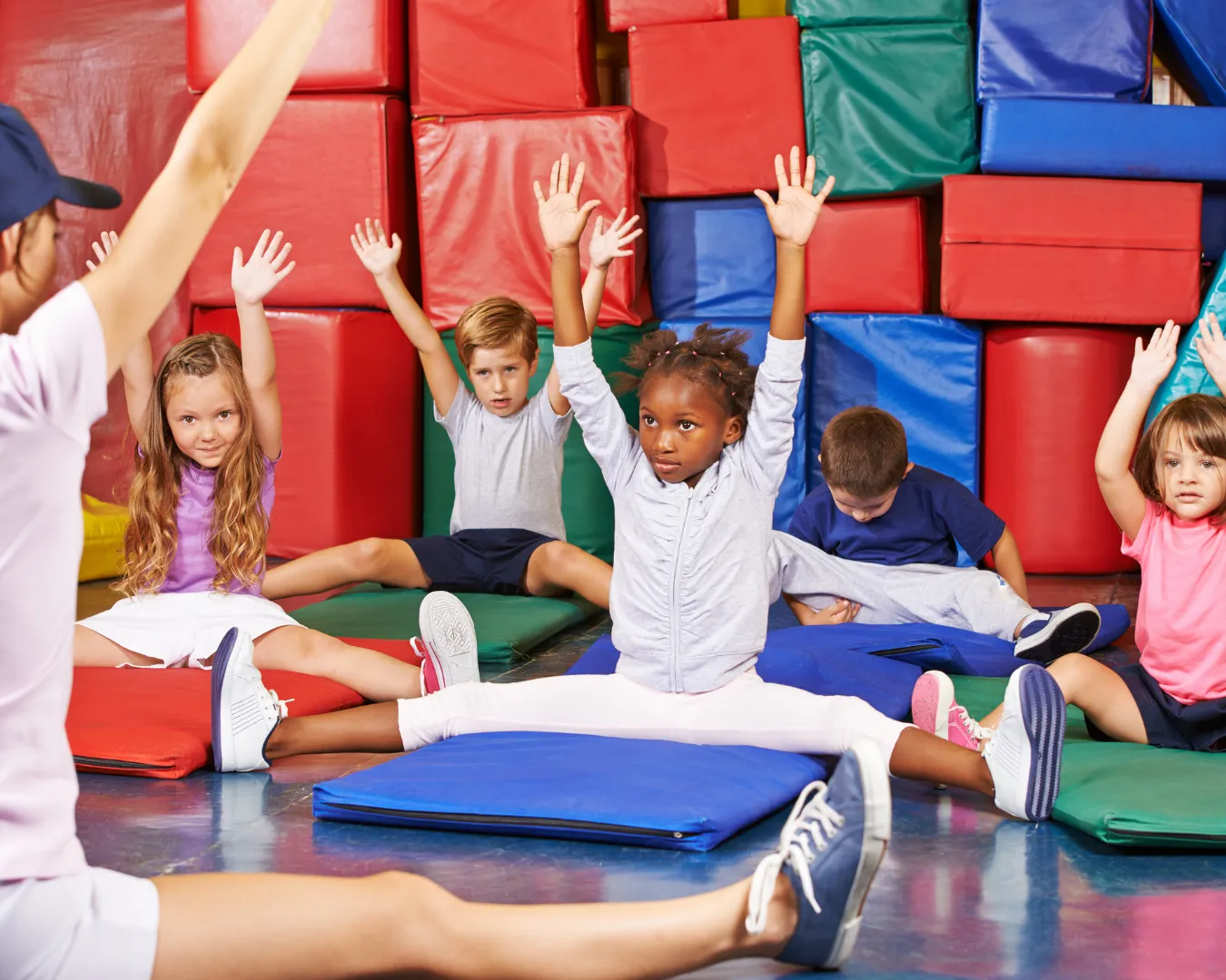 Children sitting on gym mats doing stretches with arms raised in a colorful play area.