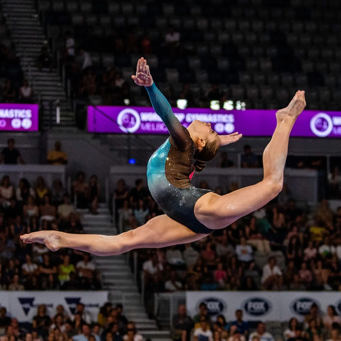 Female gymnast in a blue and black leotard performing a split leap mid-air during a competition.