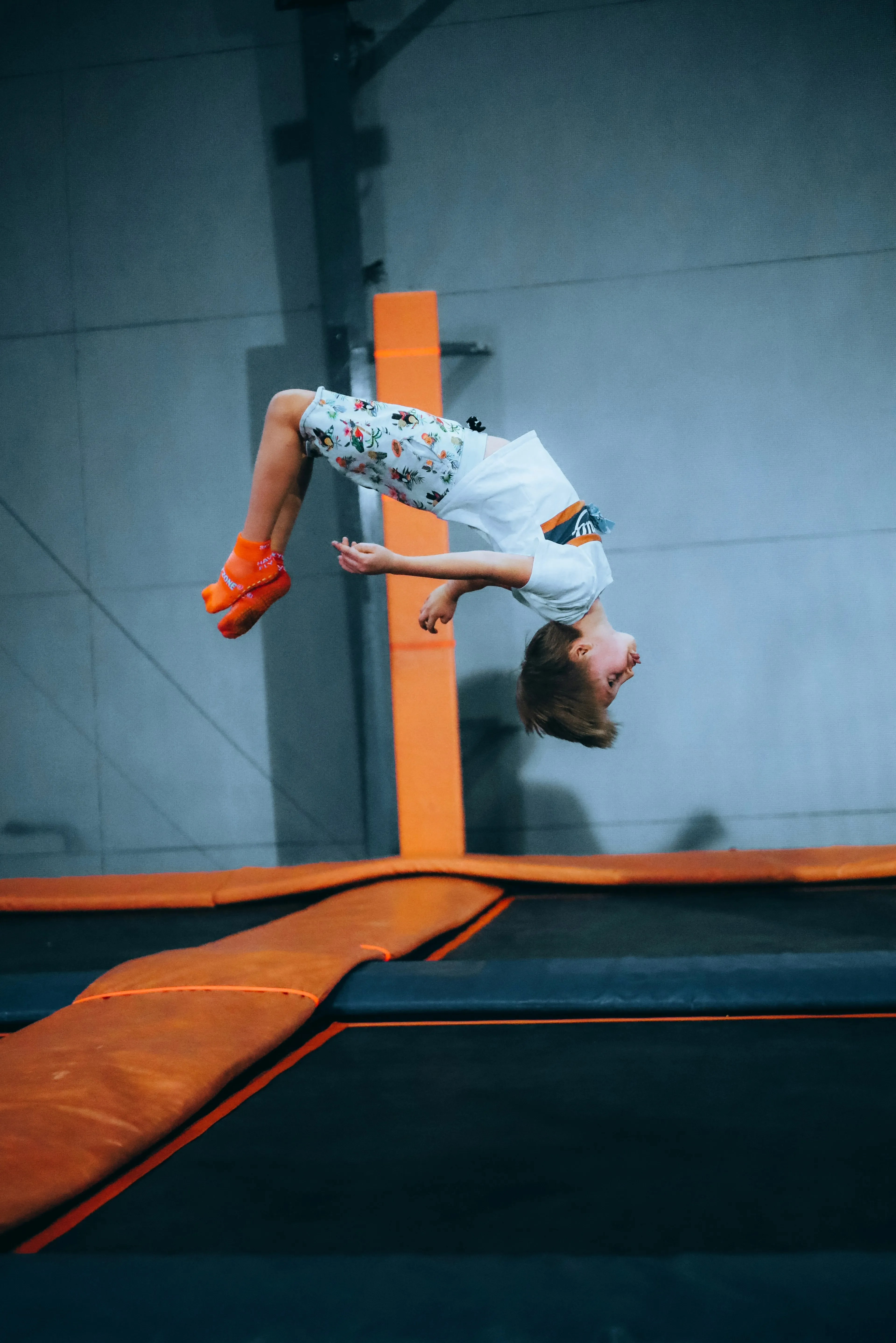 Child wearing orange socks and patterned shorts performing a backflip on an indoor trampoline.