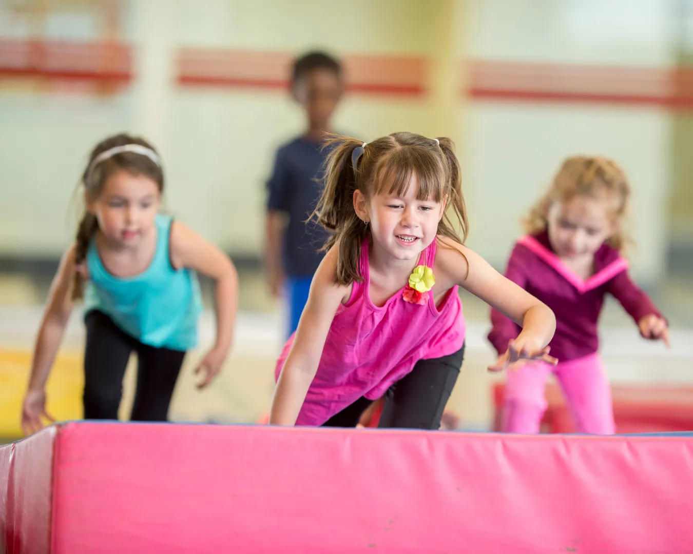 Four young children climbing over a padded gym obstacle indoors, with a girl in a pink top leading in focus.