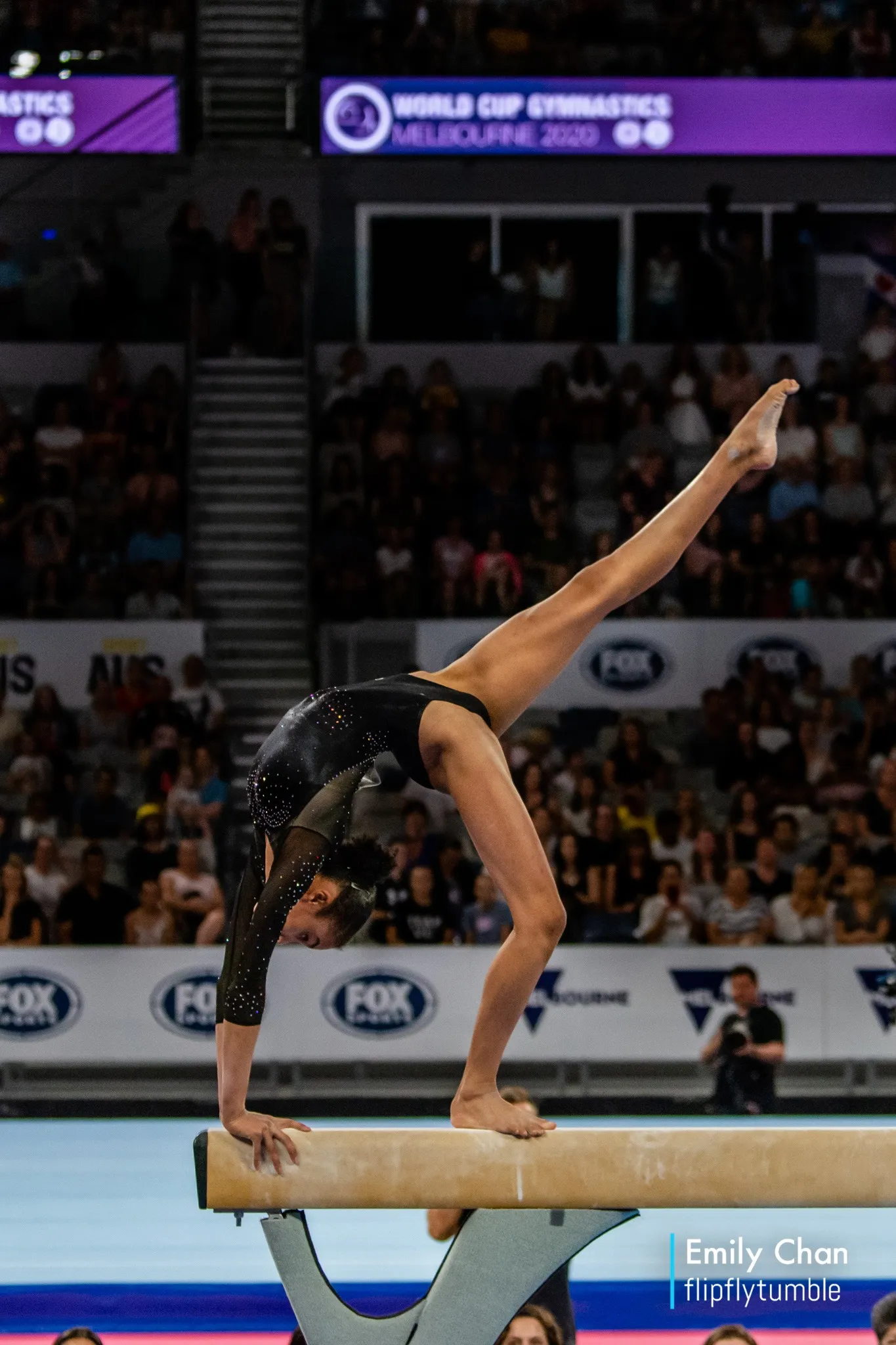 Female gymnast performing a handstand on the balance beam during the World Cup Gymnastics event with spectators in the background.