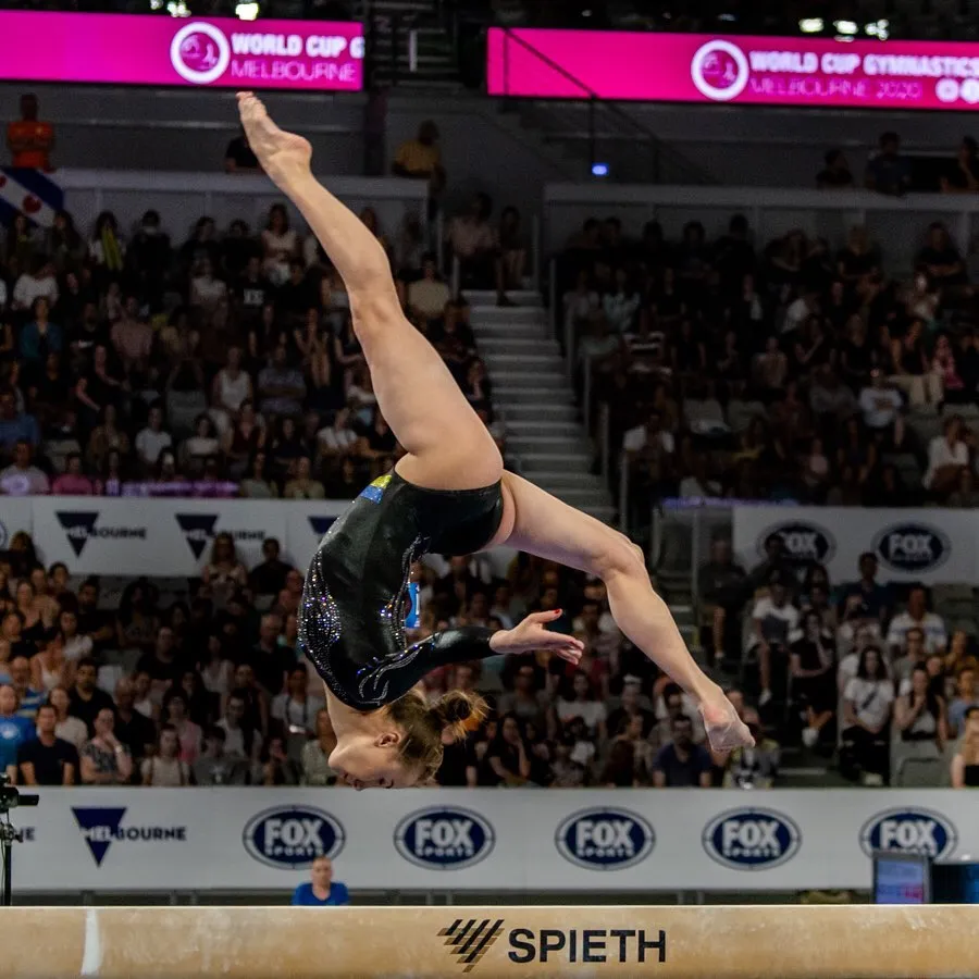 Female gymnast performing a backflip on the balance beam at a gymnastics competition with an audience watching.