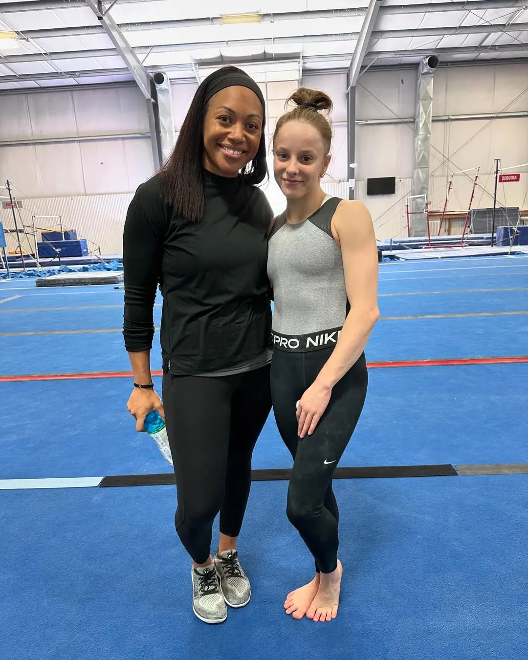 Two women standing together on a blue gymnastics floor, one in black athletic wear holding a water bottle and the other barefoot in a gray and black sleeveless top and black leggings.
