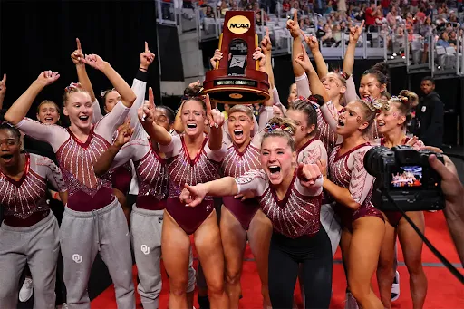Women’s gymnastics team celebrating holding up an NCAA championship trophy indoors.