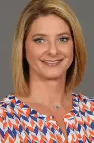 Smiling woman with blonde hair wearing a patterned blouse with blue, orange, and white colors against a gray background.