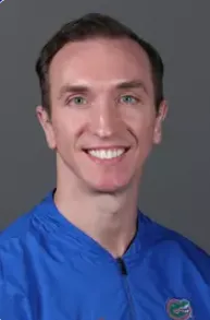 Smiling young man with short brown hair wearing a blue jacket against a gray background.